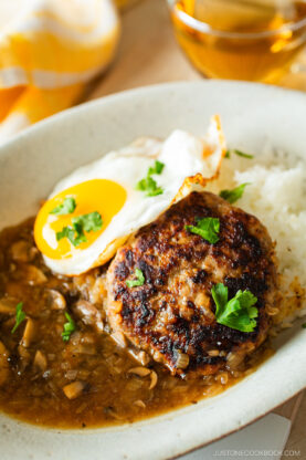 A plate with a hamburger steak topped with brown mushroom gravy, a sunny-side-up egg, white rice, and garnished with parsley. A yellow napkin and a drink are in the background.